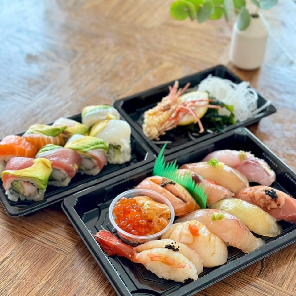 Three black trays of Hachi sushi and omakase, including nigiri, rolls, and tempura, on a wooden table.
