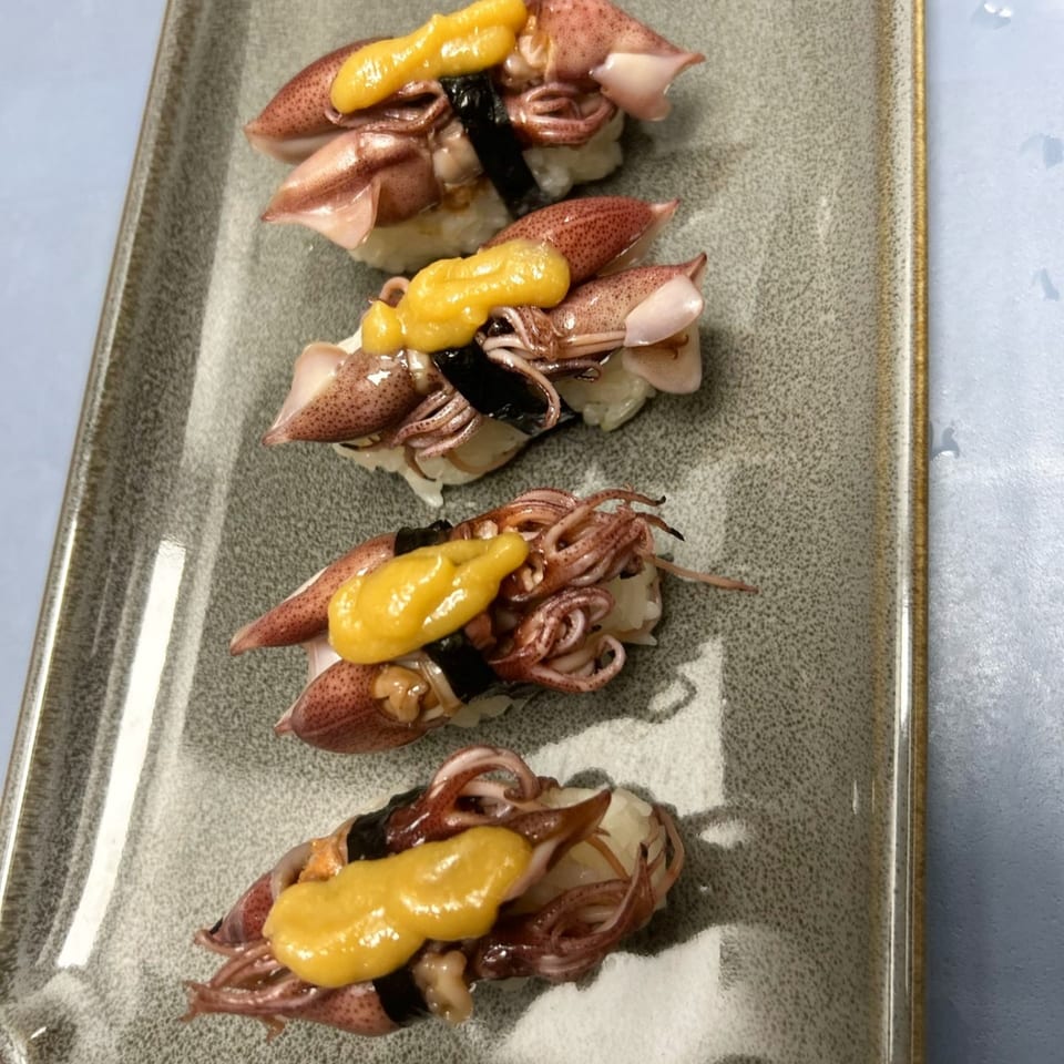 Four pieces of Hachi sushi with squid and yellow topping on a gray rectangular plate.